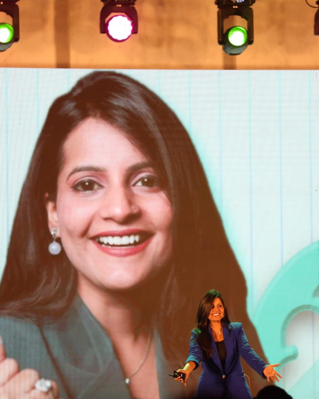 Tripti Sharma on stage, arms open, with her portrait on the giant keynote screen behind her