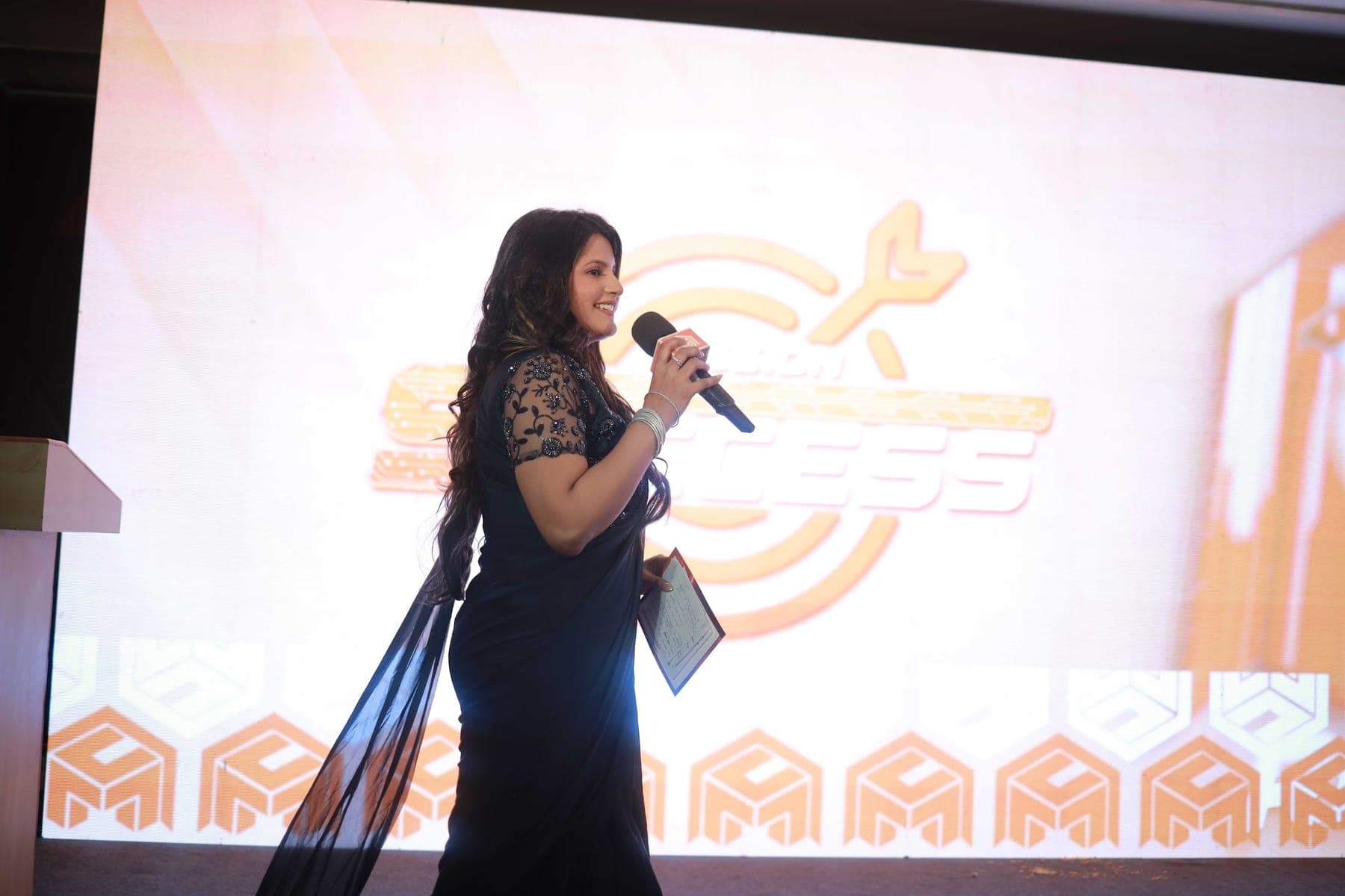 Tripti speaking with a microphone in front of the event screen